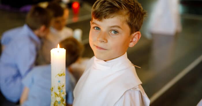 little kid boy receiving his first holy communion. happy child holding christening candle. tradition in catholic curch. kid in a white traditional gown in a church near altar.